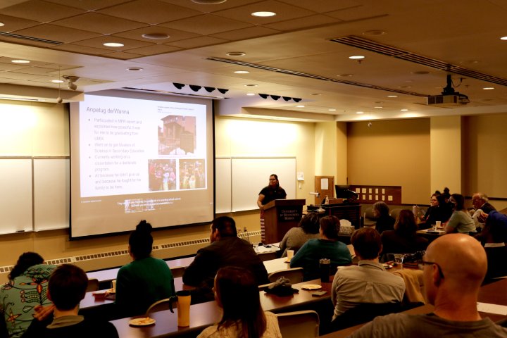 A speaker stands at a podium in front of a projected presentation in a lecture hall. The audience, seated at tiered desks, listens attentively. The screen displays a slide titled 'Anpetug de/Wanna' with text and images, while the room is softly lit. The environment appears to be educational and formal.